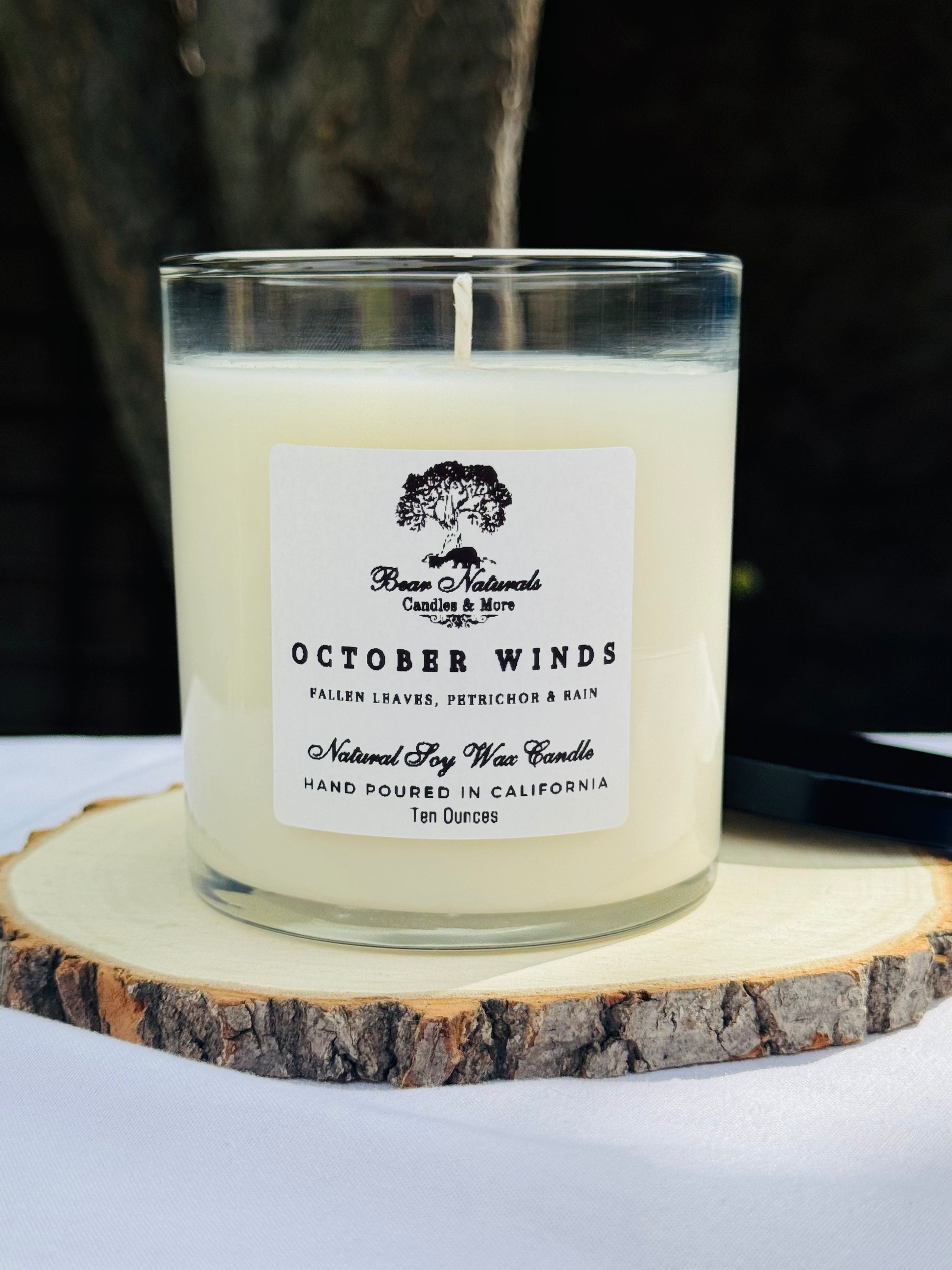 A white ten ounce candle in a clear glass jar with a black metal lid and white label with black writing sitting on a wooden surface. The candle is called "October Winds".