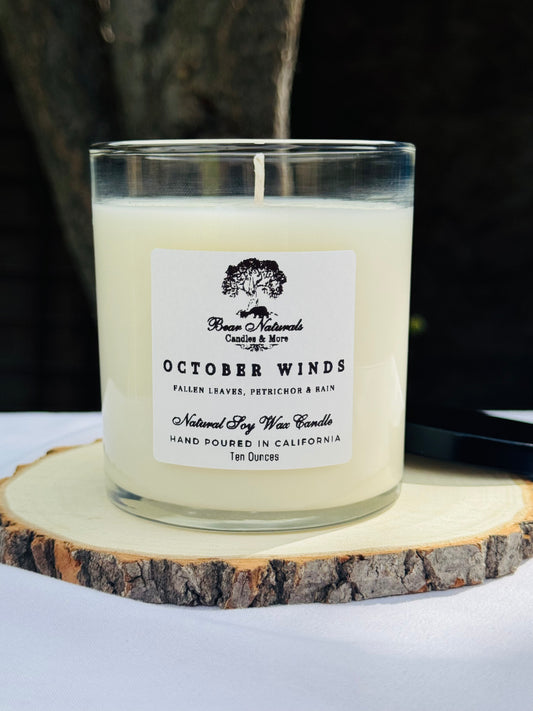 A white ten ounce candle in a clear glass jar with a black metal lid and white label with black writing sitting on a wooden surface. The candle is called "October Winds".