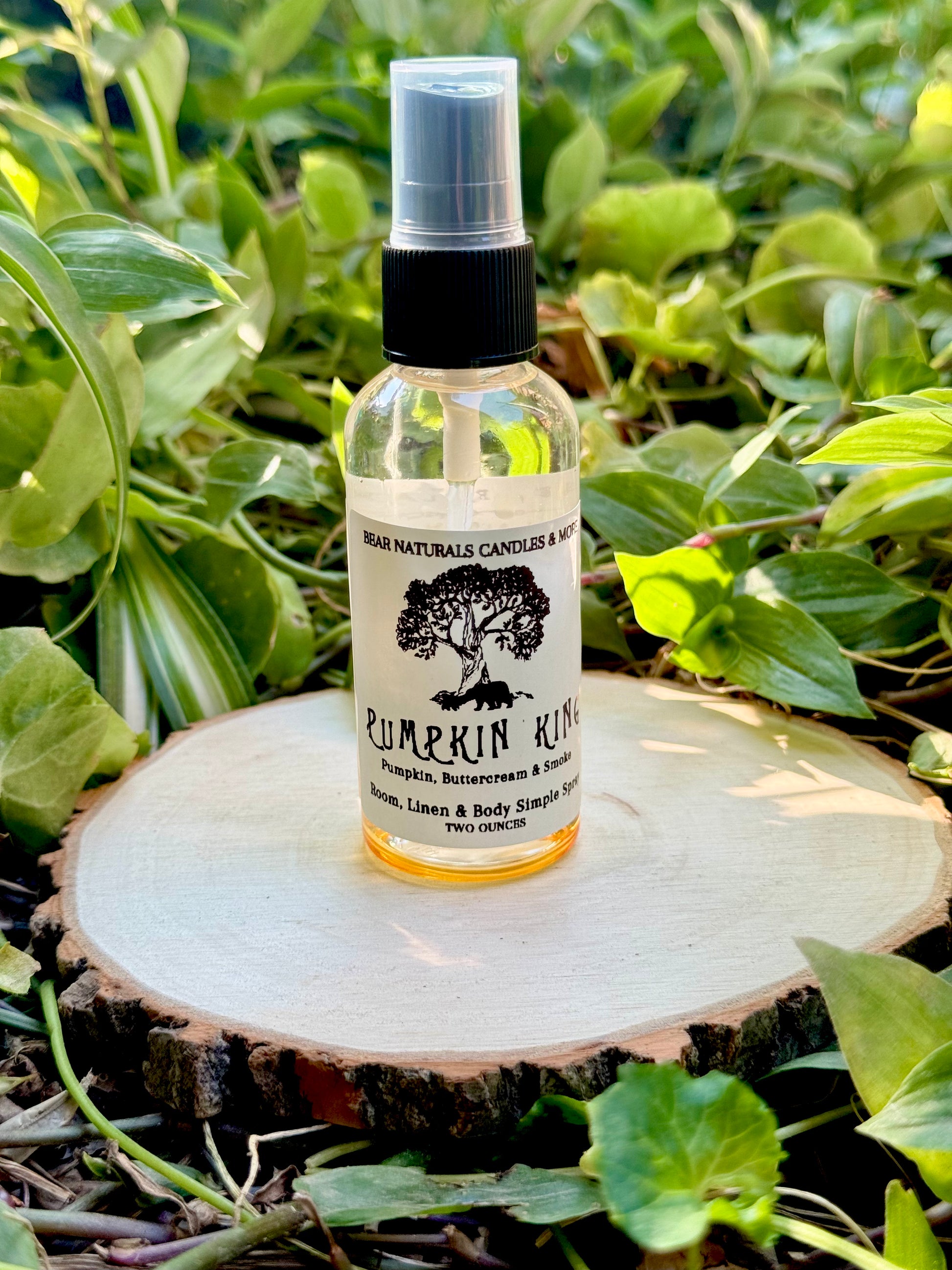 A clear two ounce finger-tip spray bottle with black and white label on a wooden surface with green leaves in the background. The bottle contains the scent "Pumpkin King"