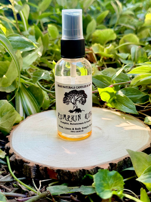 A clear two ounce finger-tip spray bottle with black and white label on a wooden surface with green leaves in the background. The bottle contains the scent "Pumpkin King"