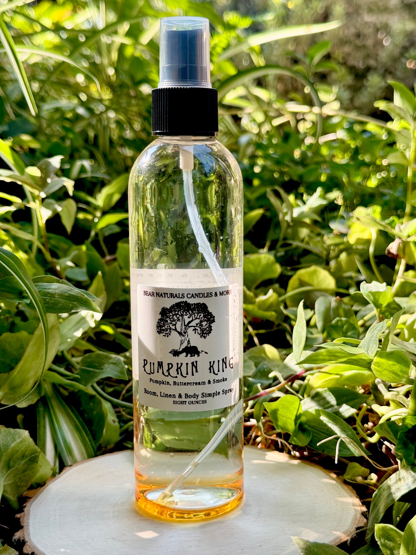 A clear eight ounce finger-tip spray bottle with black and white label on a wooden surface with green leaves in the background. The bottle contains the scent "Pumpkin King"