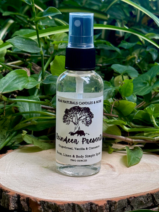 A clear two ounce finger-tip spray bottle with black and white label on a wooden surface with green leaves in the background. The bottle contains the scent "Reindeer Presents".
