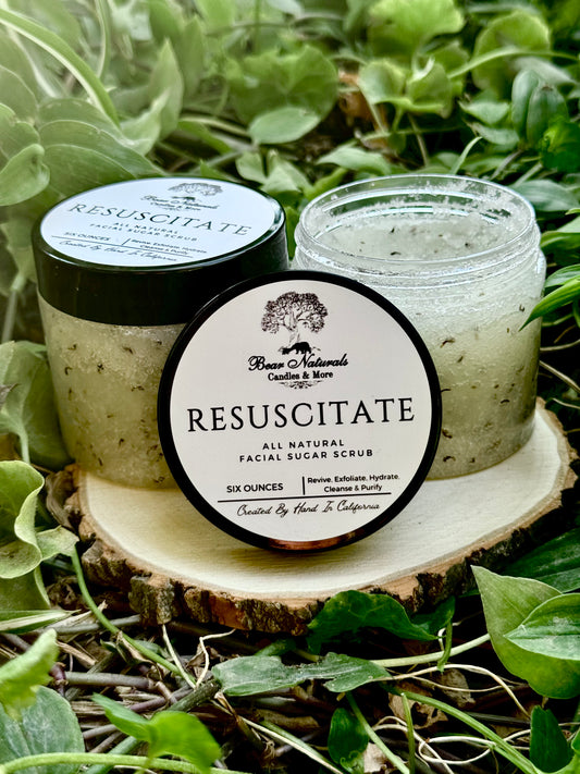Two clear six ounce plastic jars with black lids featuring a white label with black writing that reads "Resuscitate All Natural Facial Sugar Scrub" filled with product sitting on a wooden base with green leaves in the background..