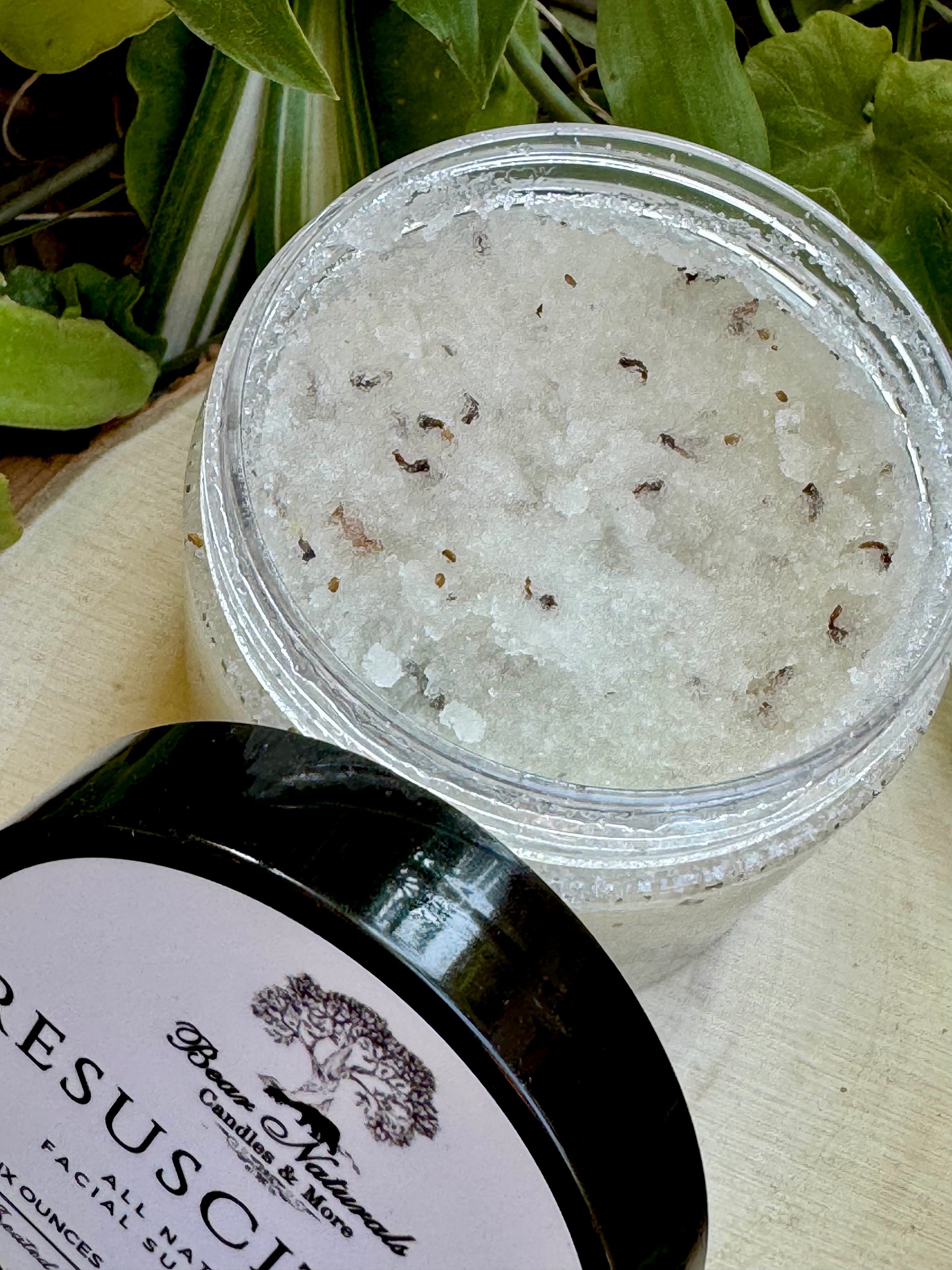 Jar of Resuscitate All Natural Facial scrub with a black lid on a wooden surface with green leaves in the background.
