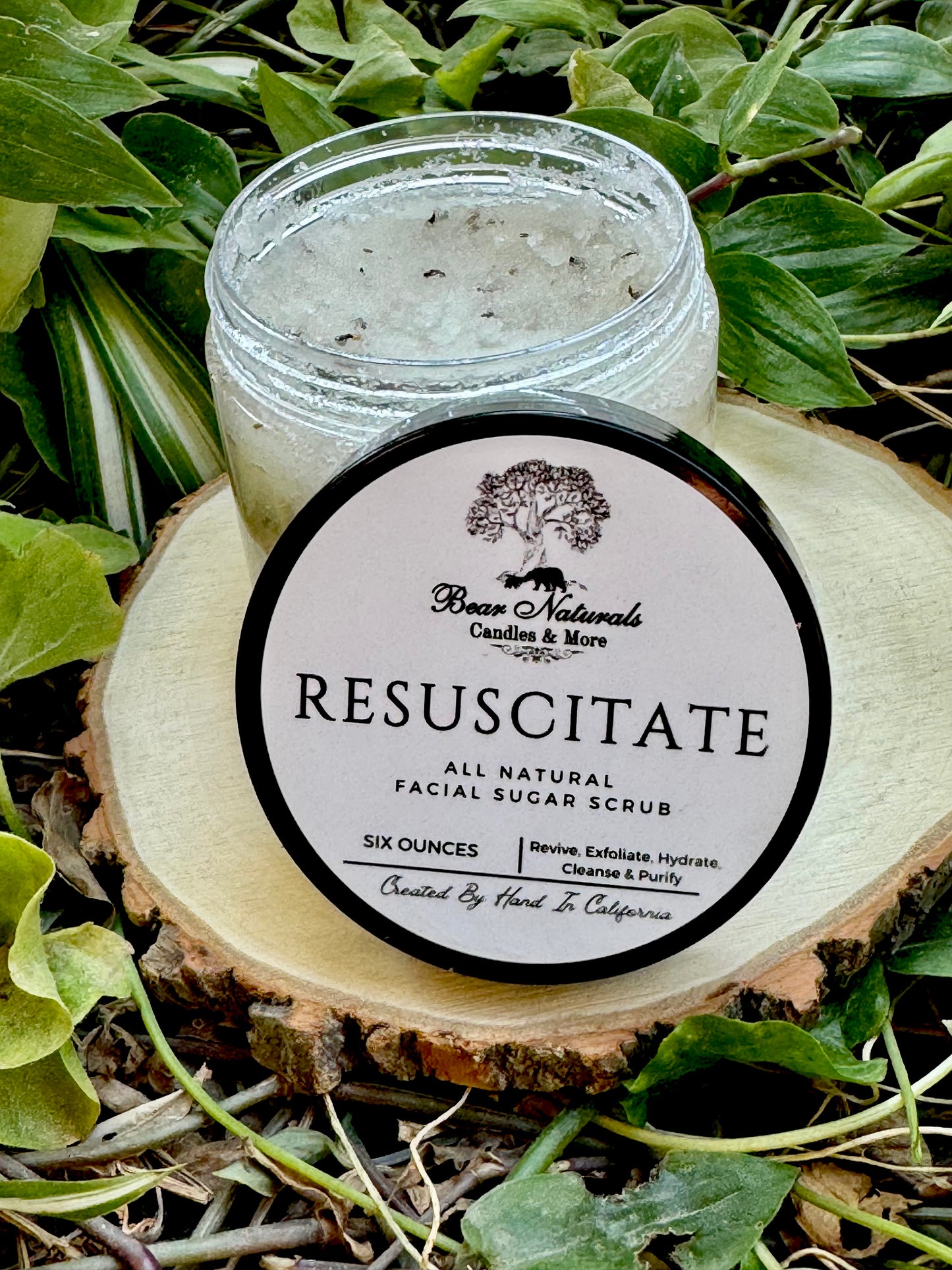 Jar of Resuscitate All Natural Facial scrub with a black lid on a wooden surface with green leaves in the background.