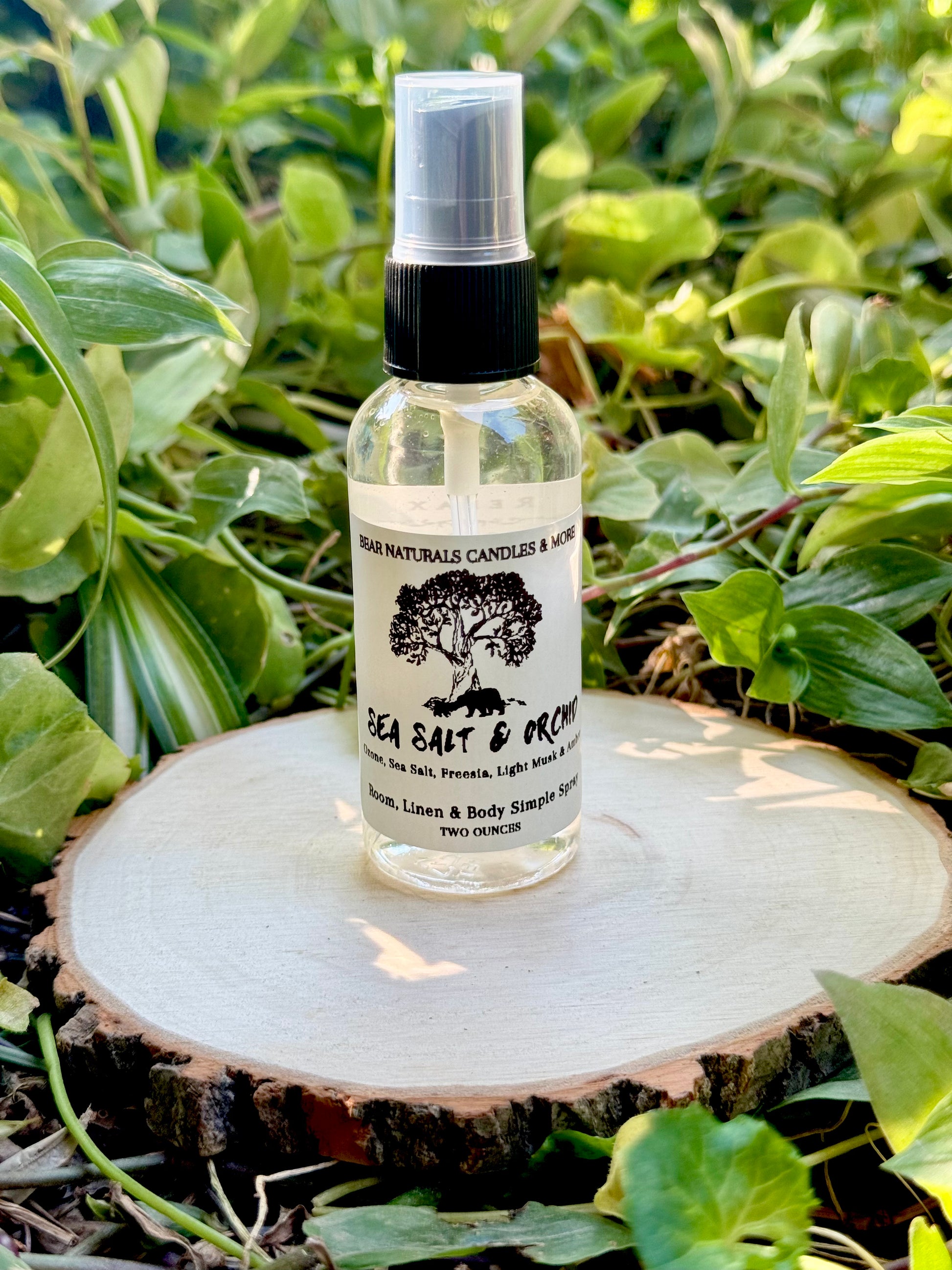 A clear two ounce finger-tip spray bottle with black and white label on a wooden surface with green leaves in the background. The bottle contains the scent "Sea Salt And Orchid".
