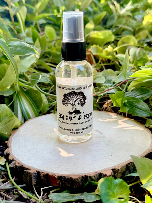 A clear two ounce finger-tip spray bottle with black and white label on a wooden surface with green leaves in the background. The bottle contains the scent "Sea Salt And Orchid".
