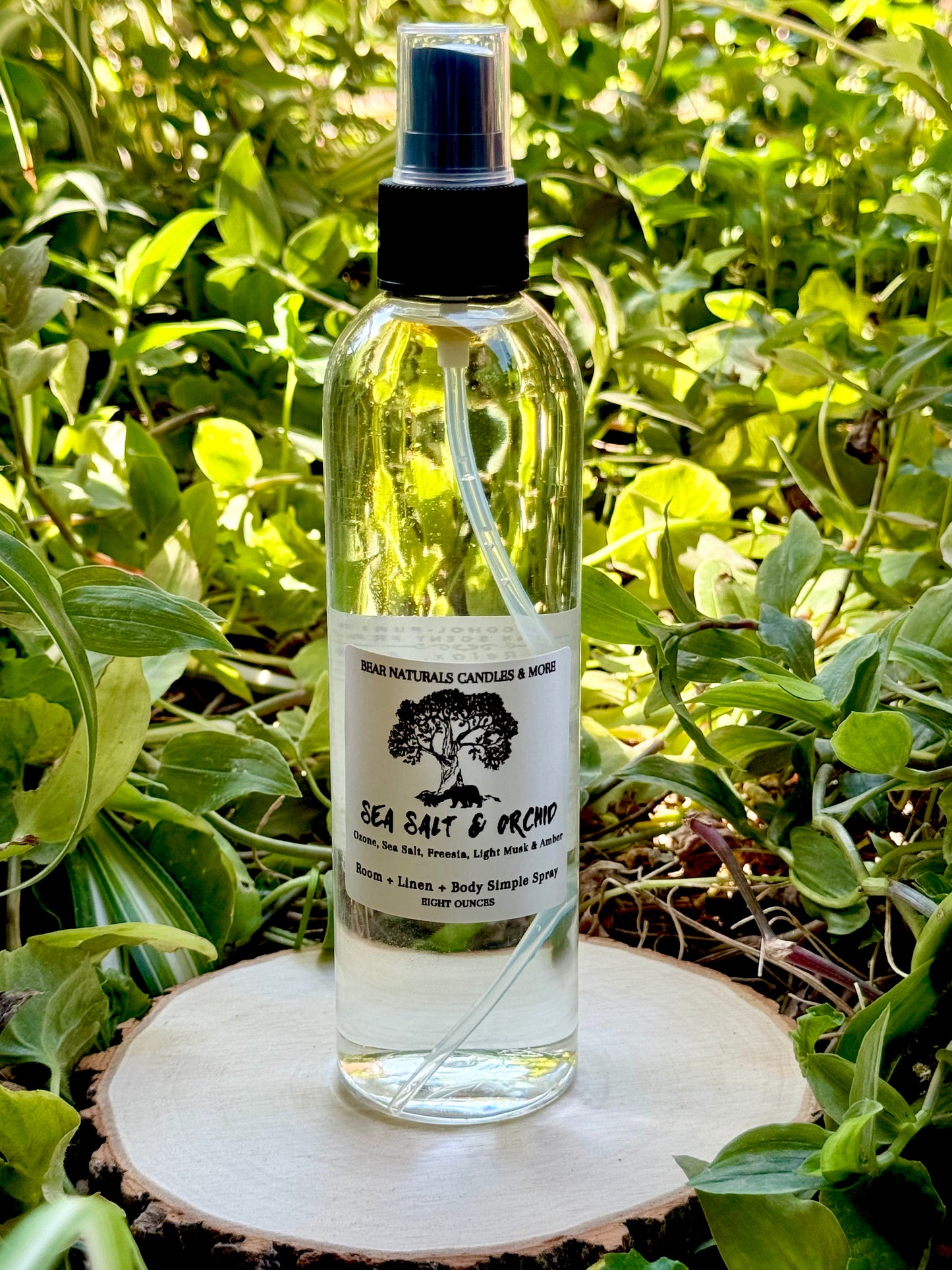 A clear eight ounce finger-tip spray bottle with black and white label on a wooden surface with green leaves in the background. The bottle contains the scent "Sea Salt And Orchid".