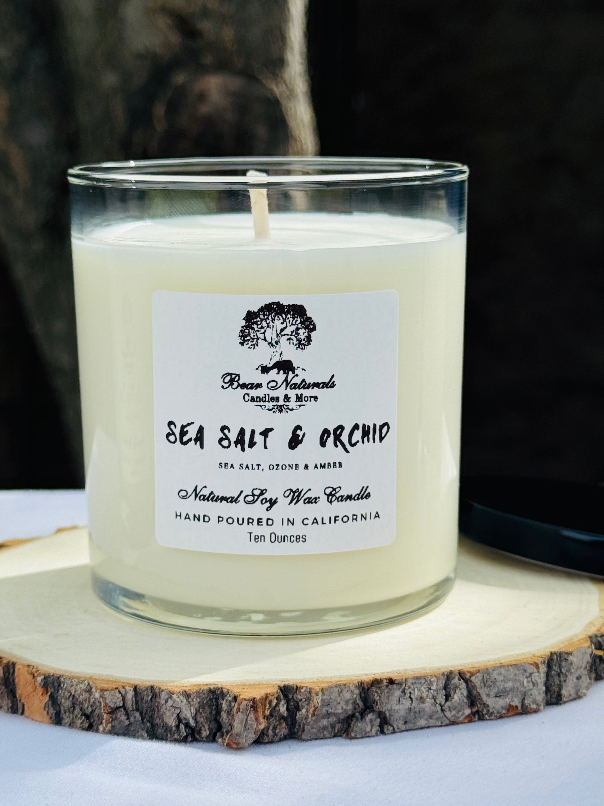 A white ten ounce candle in a clear glass jar with a black metal lid and white label with black writing sitting on a wooden surface. The candle is called "Sea Salt & Orchid".