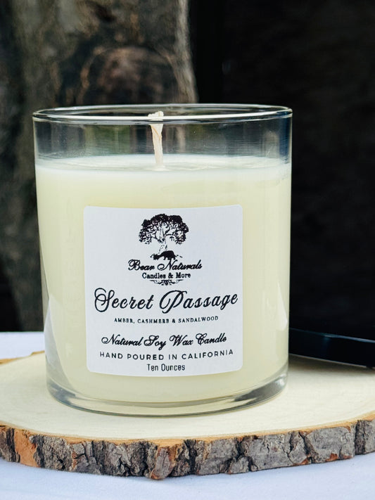 A white ten ounce candle in a clear glass jar with a black metal lid and white label with black writing sitting on a wooden surface. The candle is called "Secret Passage".