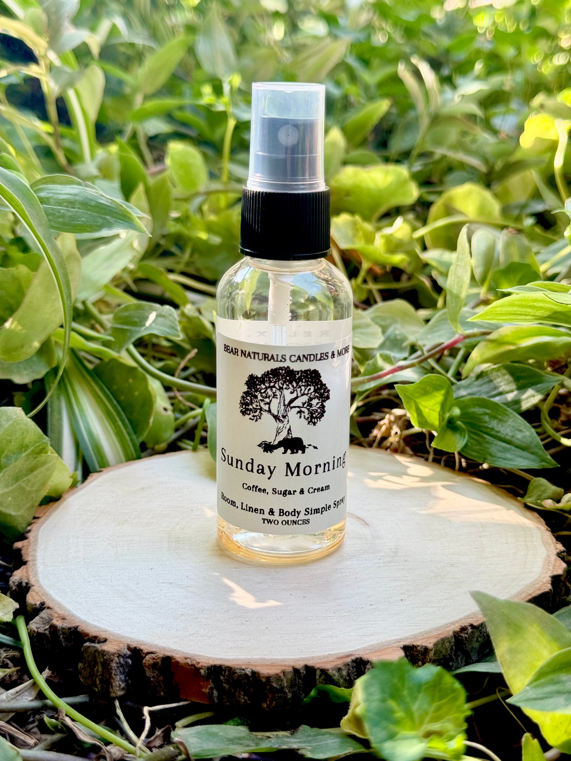 A clear two ounce finger-tip spray bottle with black and white label on a wooden surface with green leaves in the background. The bottle contains the scent "Sunday Morning".