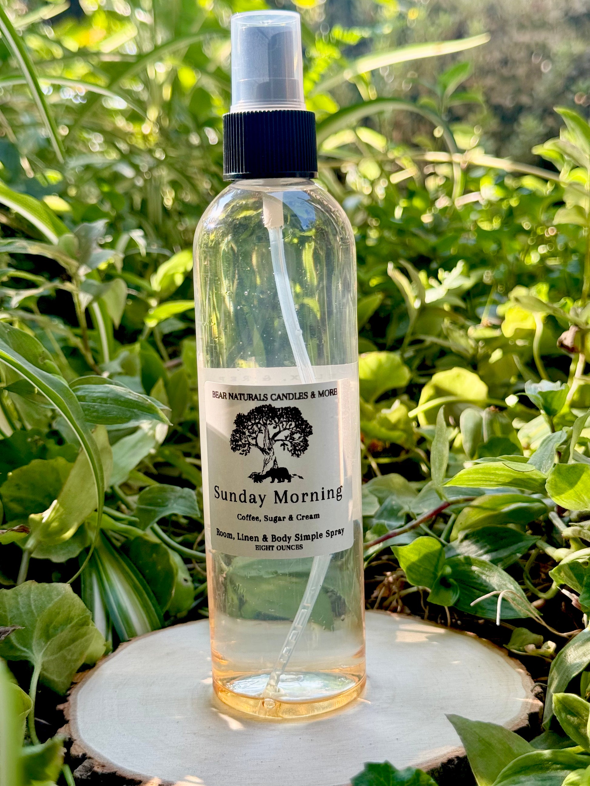 A clear eight ounce finger-tip spray bottle with black and white label on a wooden surface with green leaves in the background. The bottle contains the scent "Sunday Morning".