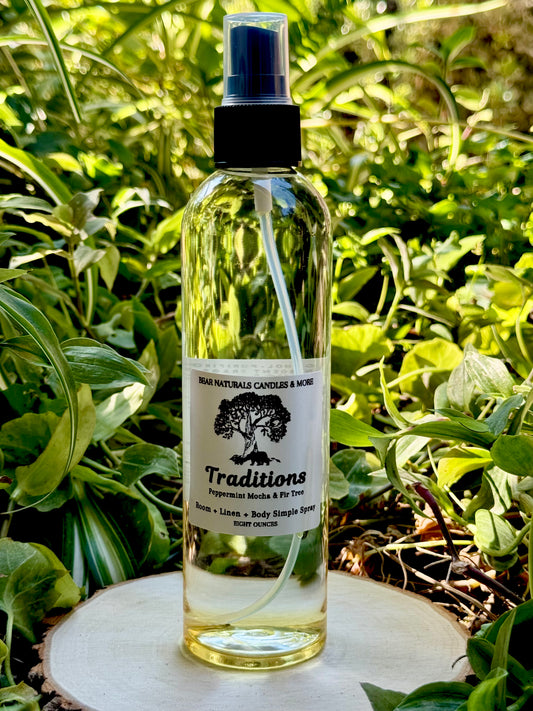 A clear eight ounce finger-tip spray bottle with black and white label on a wooden surface with green leaves in the background. The bottle contains the scent "Traditions".
